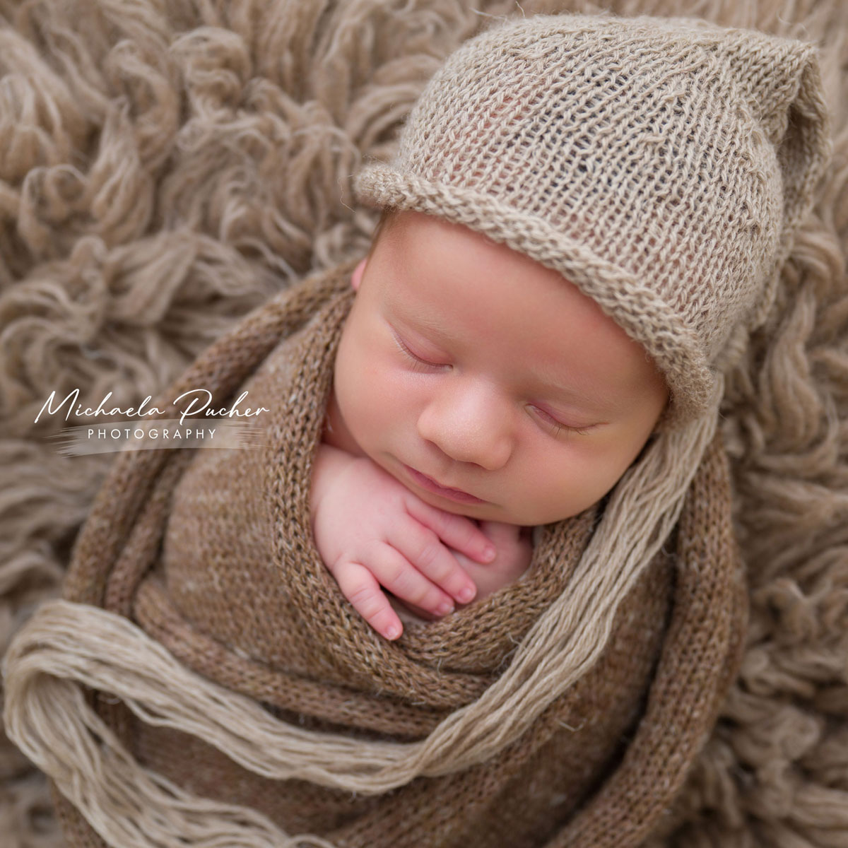 Schlafendes Neugeborenes, eingewickelt in braune Stricktücher, mit einer gestrickten Zipfelmütze, liegend auf einer flauschigen Unterlage.